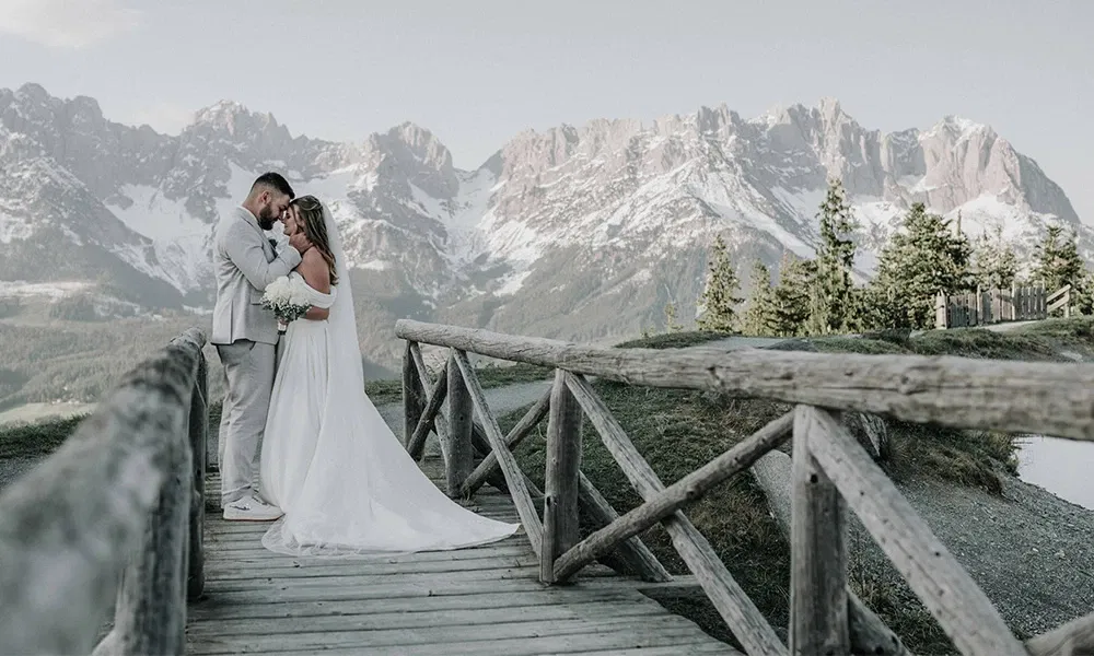 Brautpaar auf Holzbrücke mit Blick auf verschneite Bergkulisse, verliebt aneinander gelehnt.