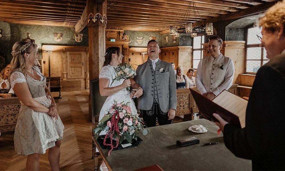 Fröhliches Brautpaar in Tracht lacht laut während der Trauung im rustikalen Standesamt.