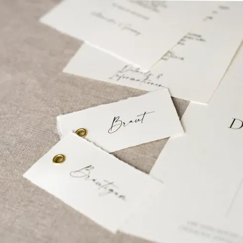 Feine Hochzeitspapeterie mit handbeschrifteten Kärtchen für Braut und Bräutigam, stilvoll arrangiert auf Leinenstoff.