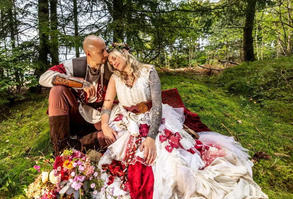 Braut und Bräutigam im Wikinger Stil sitzen eng umschlungen auf moosigem Waldboden mit Brautstrauß