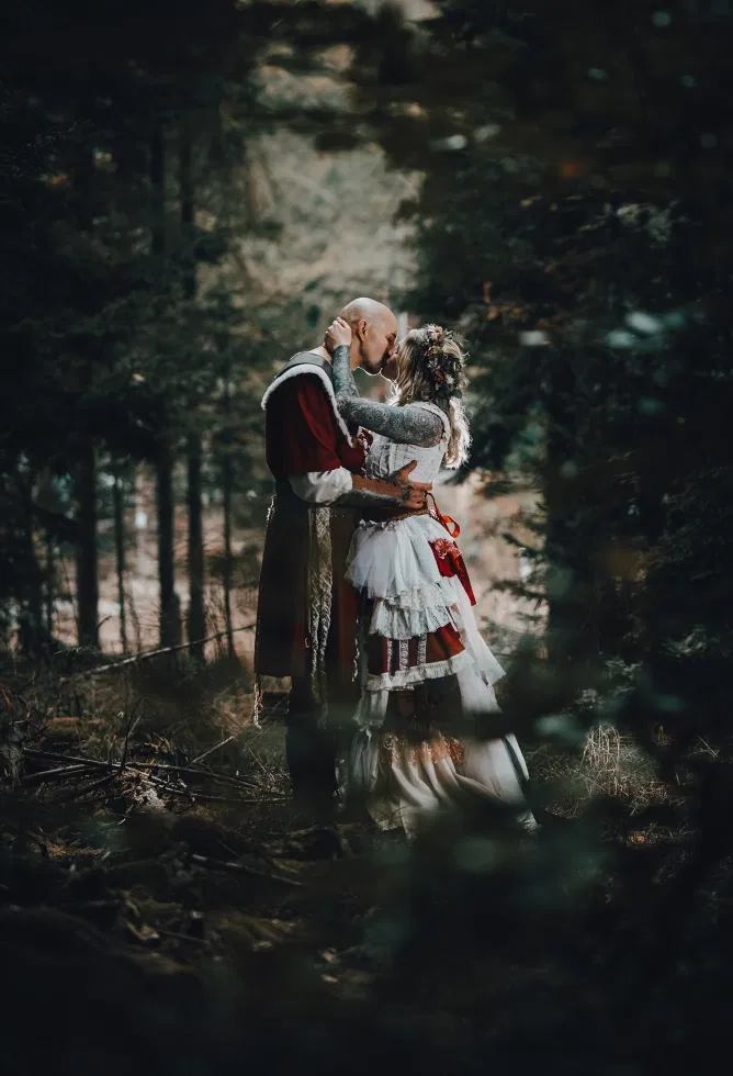 Romantisches Brautpaar im Wikinger Look küsst sich innig im dunklen Wald zwischen hohen Bäumen
