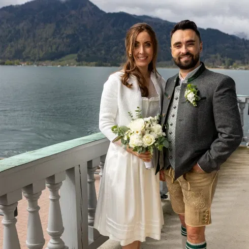 Braut und Bräutigam im Trachten-Outfit bei ihrer Hochzeit am Tegernsee, mit Blick auf den See und die umliegenden Berge.