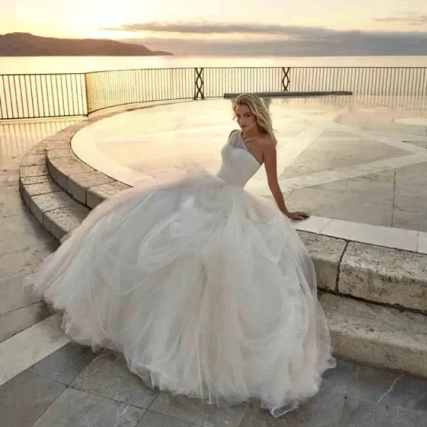 Braut in opulentem Tüllkleid mit asymmetrischem Träger sitzt vor romantischer Meereskulisse bei Sonnenuntergang.