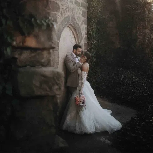 Hochzeitspaar steht innig an einer alten Steinmauer, die Braut im Spitzenkleid hält einen Blumenstrauß mit pastellfarbenen Rosen.
