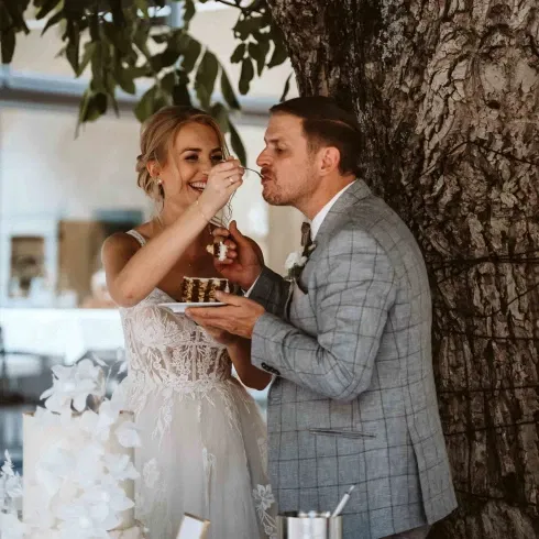 Braut füttert Bräutigam lachend mit einem Stück Hochzeitstorte unter einem Baum beim Anschneiden der Torte.