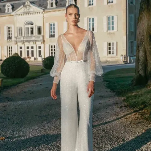 Braut in funkelndem weißen Jumpsuit mit transparentem Oberteil und Puffärmeln vor einem eleganten Schloss.