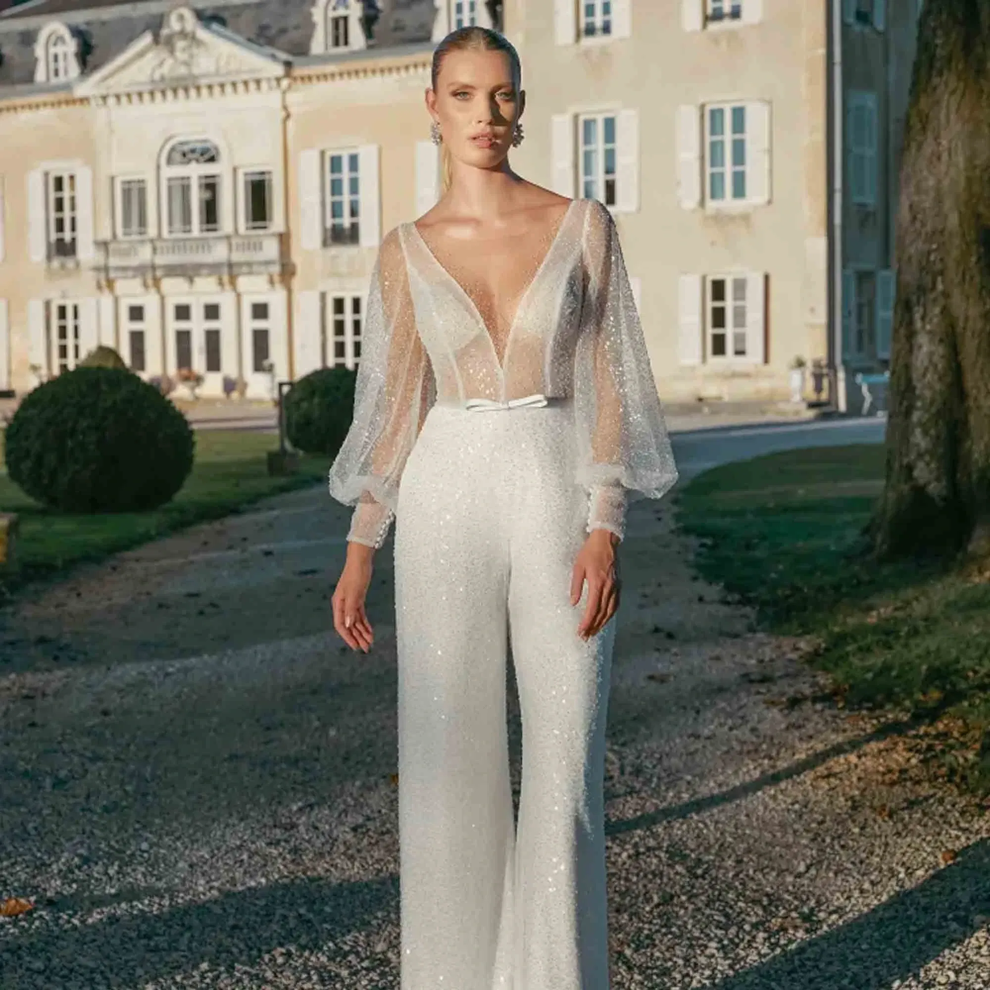 Braut in funkelndem weißen Jumpsuit mit transparentem Oberteil und Puffärmeln vor einem eleganten Schloss.