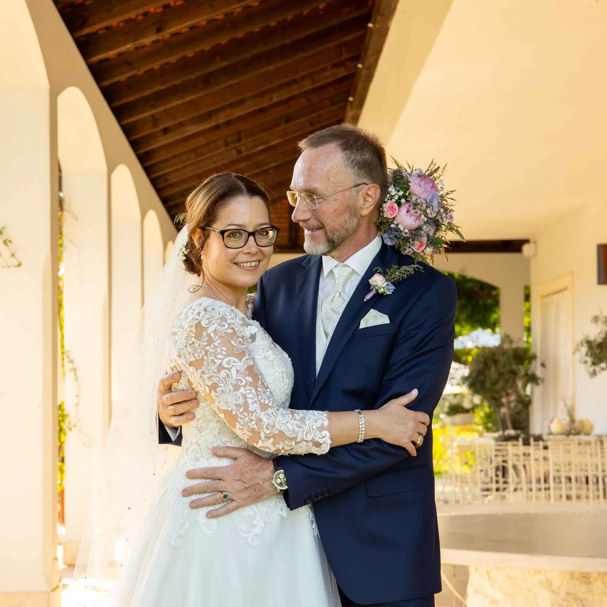 Braut und Bräutigam umarmen sich lächelnd unter einem Arkadengang, beide in festlicher Hochzeitskleidung.