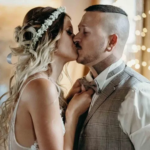 Braut mit Blumenkranz küsst ihren Bräutigam in Weste und Fliege bei einer romantischen Hochzeitsszene.