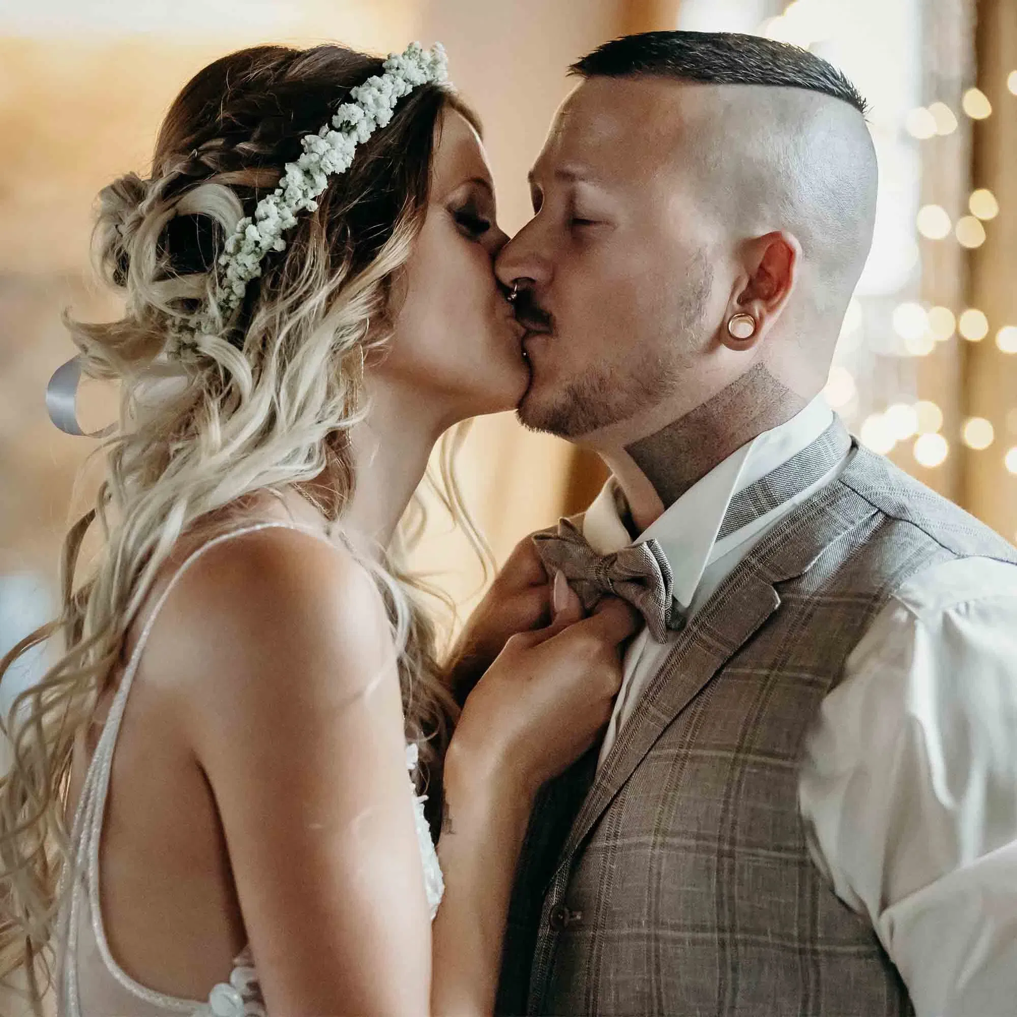 Braut mit Blumenkranz küsst ihren Bräutigam in Weste und Fliege bei einer romantischen Hochzeitsszene.