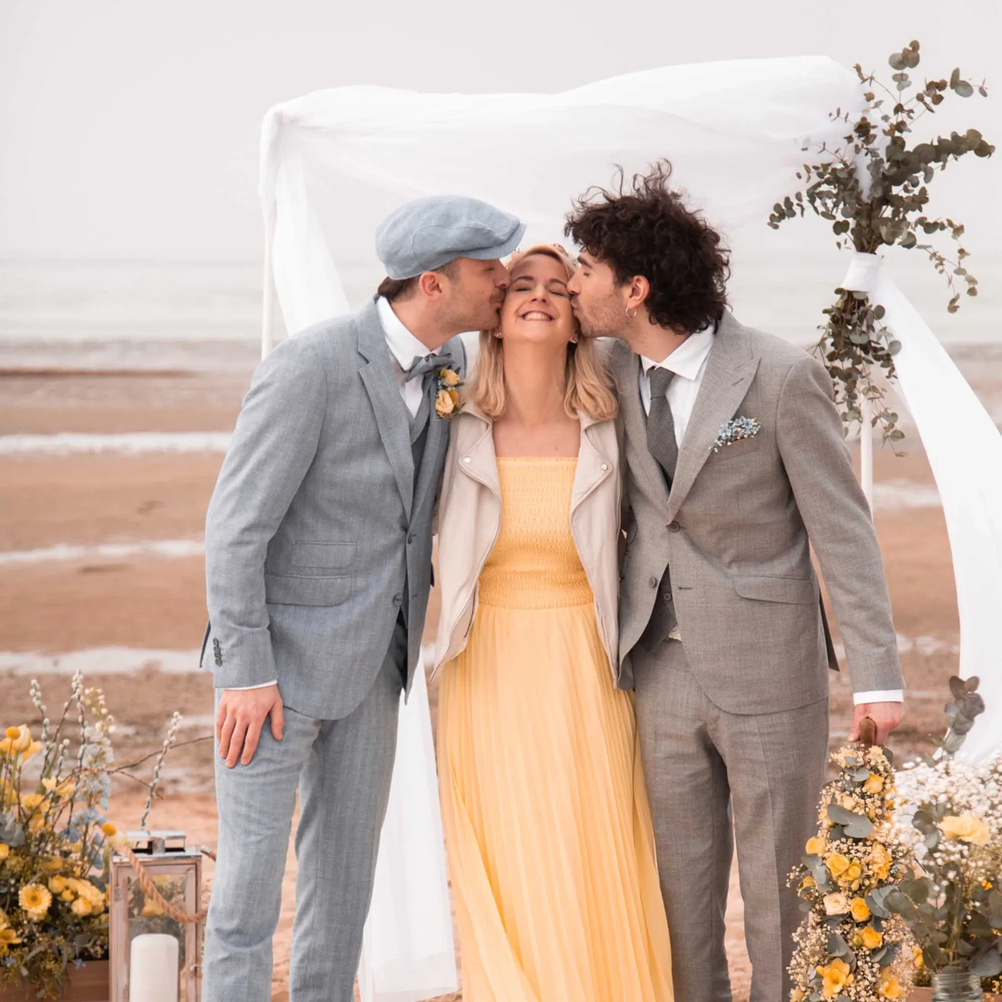 Zwei Männer küssen eine lachende Frau auf die Wangen bei einer freien Trauung am Strand, festlich gekleidet vor einem weißen Traubogen mit gelber Blumendeko.