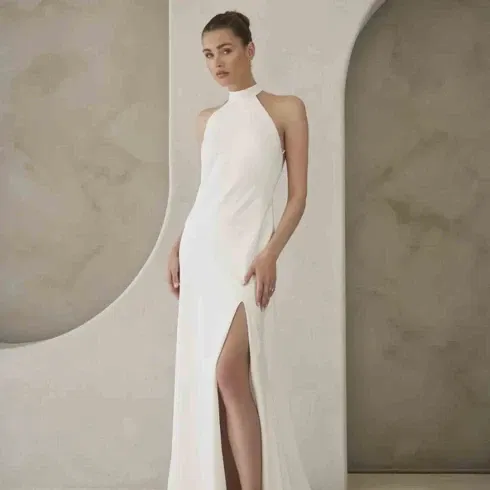 Schlichtes, ärmelloses Brautkleid mit hohem Kragen und seitlichem Beinschlitz in moderner, minimalistischer Umgebung.
