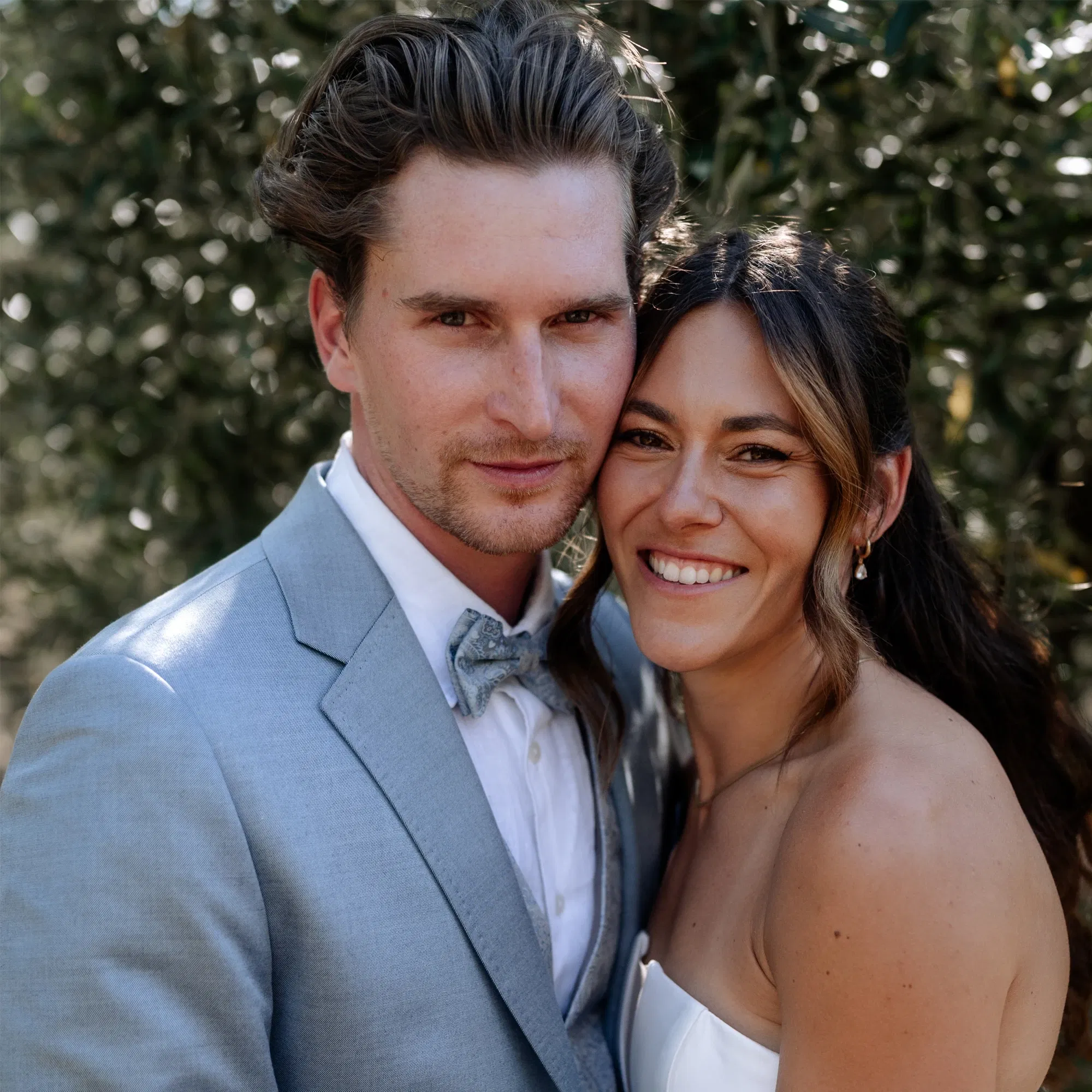 Braut und Bräutigam lächeln eng umschlungen vor Olivenbäumen in die Kamera – Sommerhochzeit mit mediterranem Flair.