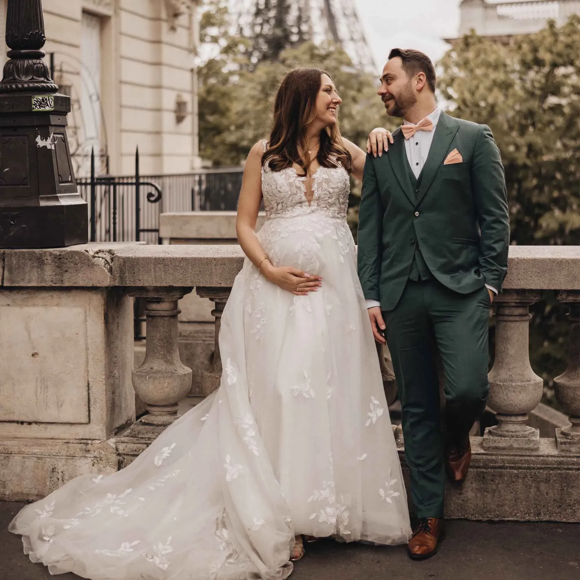 Schwangere Braut in weißem Kleid und ihr Bräutigam im grünen Anzug beim After-Wedding-Shooting in Paris mit Blick auf den Eiffelturm.