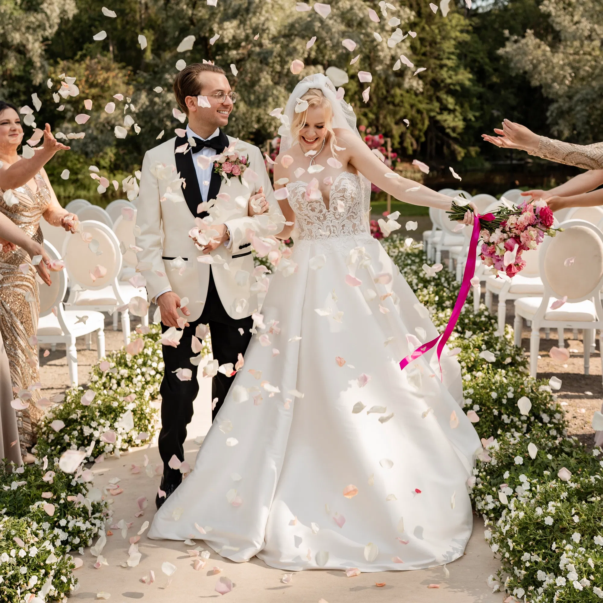 Braut und Bräutigam gehen strahlend den Blumenweg entlang, während Gäste sie mit Rosenblättern bewerfen.