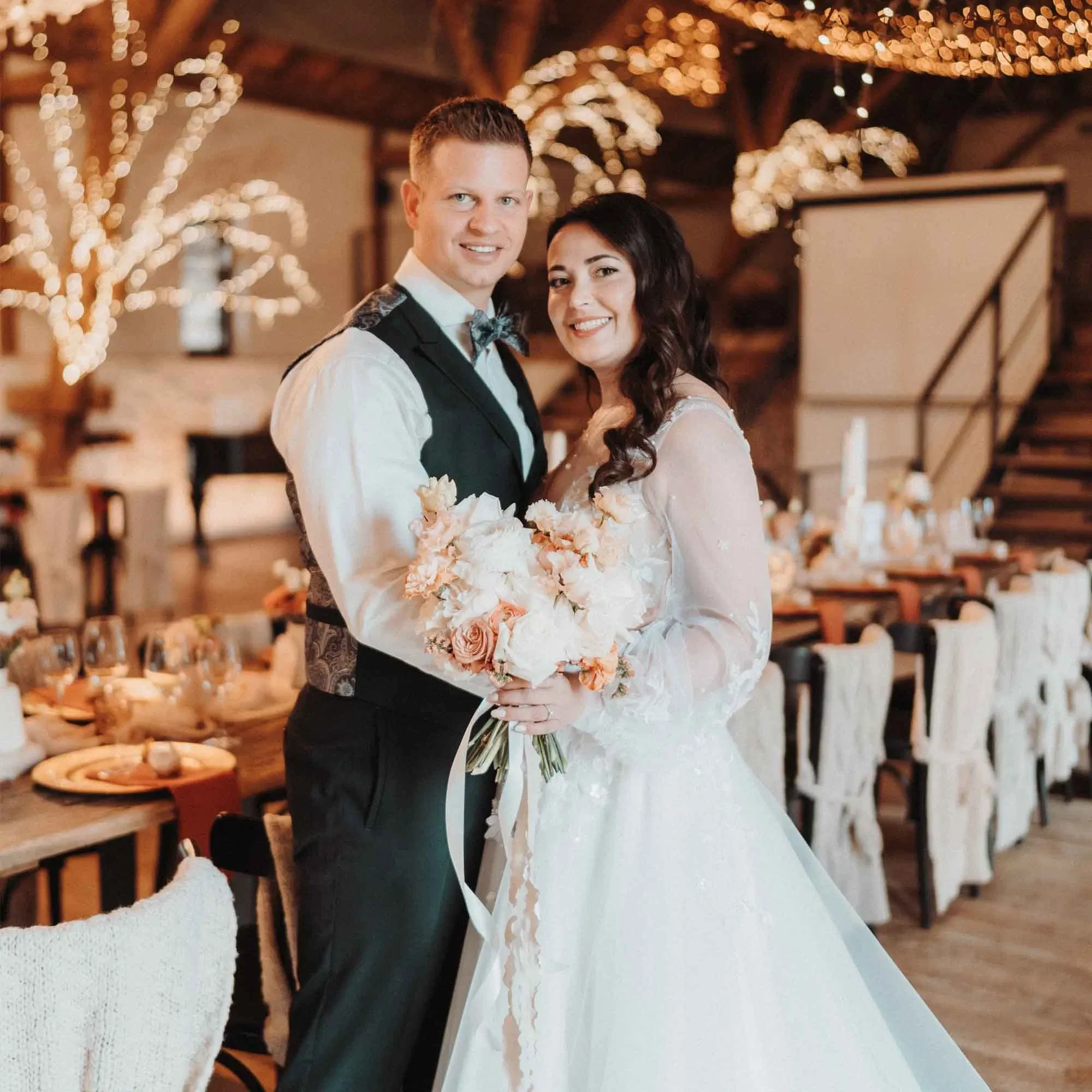 Ein Brautpaar posiert lächelnd in festlich dekorierter Scheunenlocation mit Lichterketten und festlicher Tischdekoration.