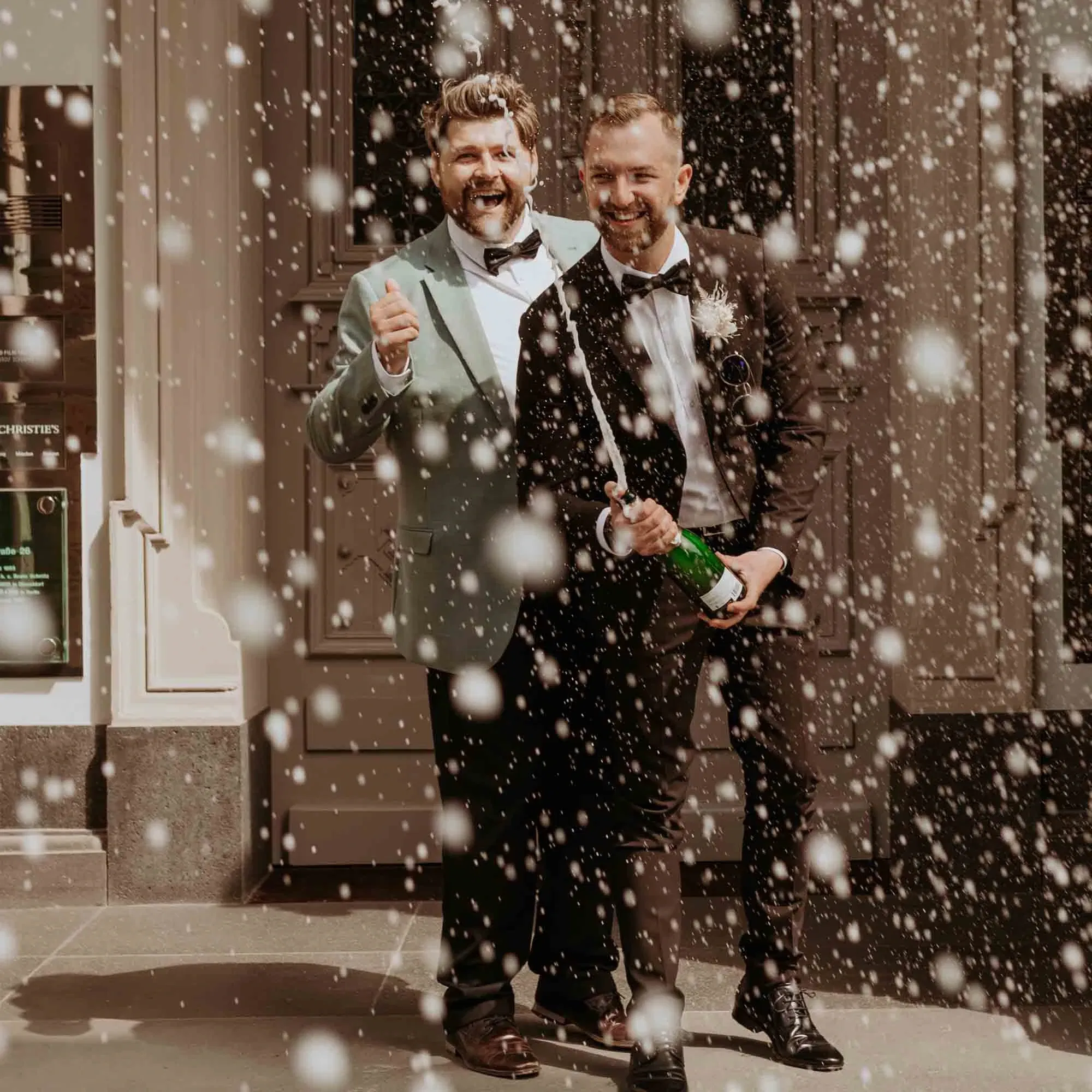 Zwei Bräutigame im Smoking feiern vor einer Location, während Champagner sprudelt und Schaum in der Luft tanzt.
