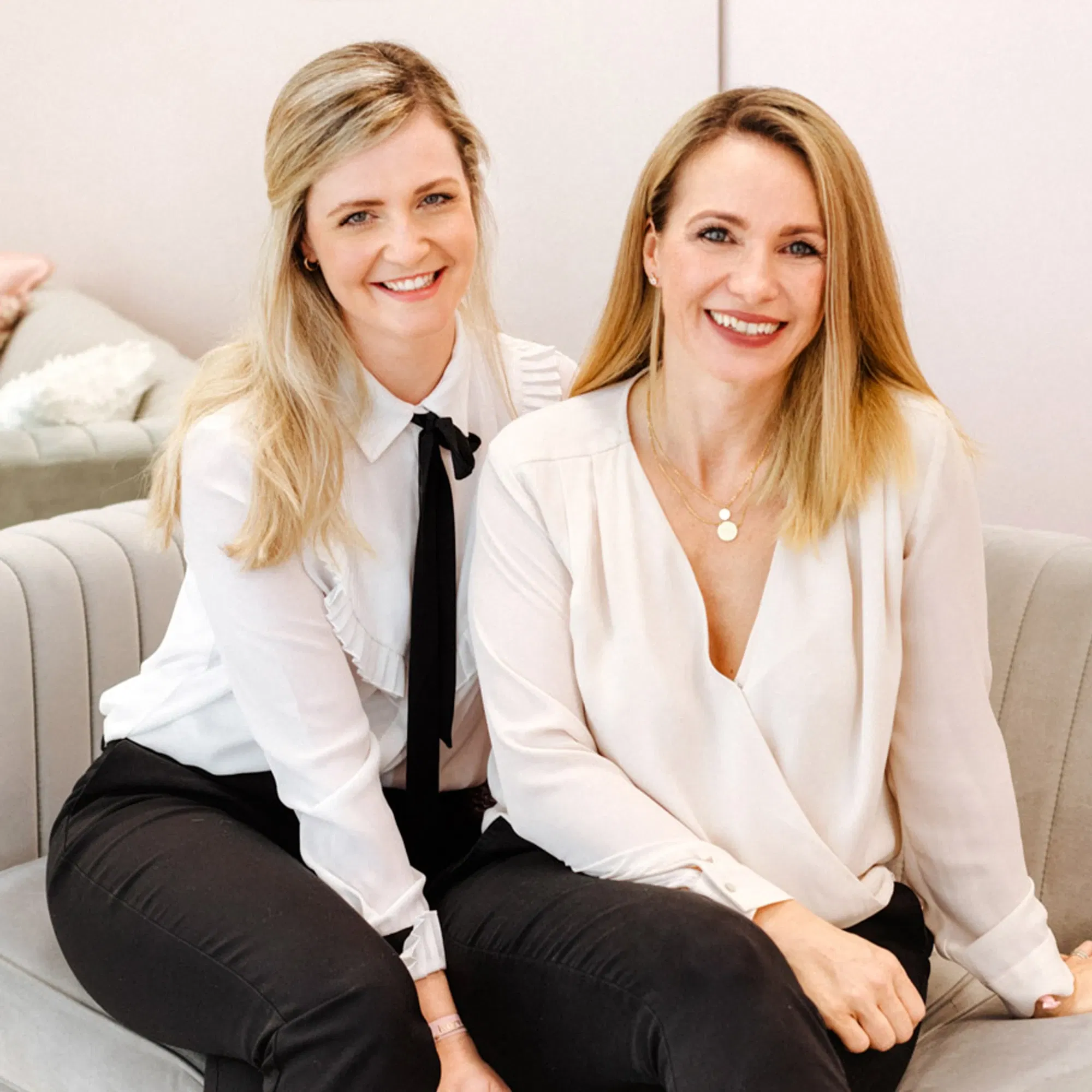 Portrait von zwei Frauen in heller Bluse und dunkler Hose, die gemeinsam auf einem Sofa posieren.