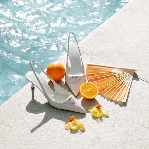 Weiße Prada-High-Heels am Pool, dekoriert mit einer Orange, einem Fächer und gelben Blüten für sommerliche Hochzeitsinspiration.