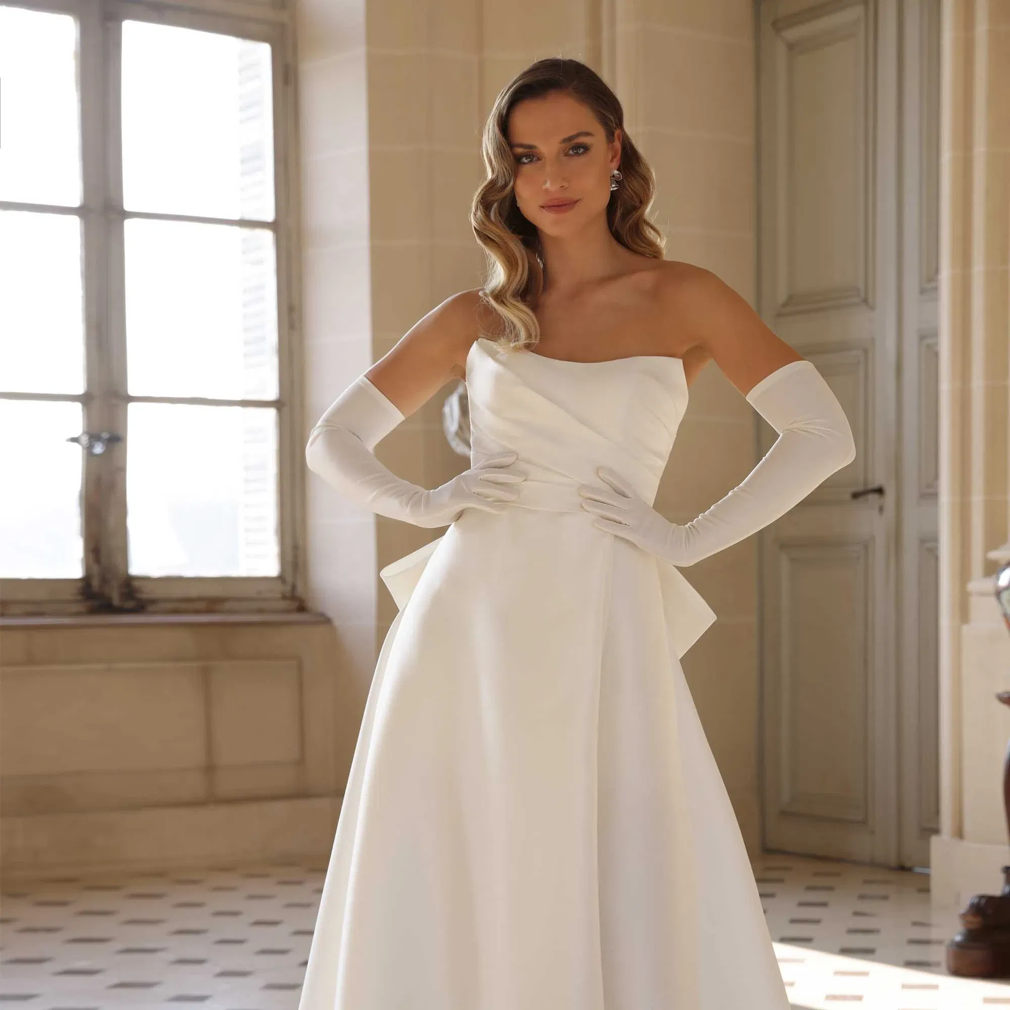 Elegante Braut in einem schulterfreien Satin-Brautkleid mit langen Handschuhen posiert in einem hellen Schlossraum.