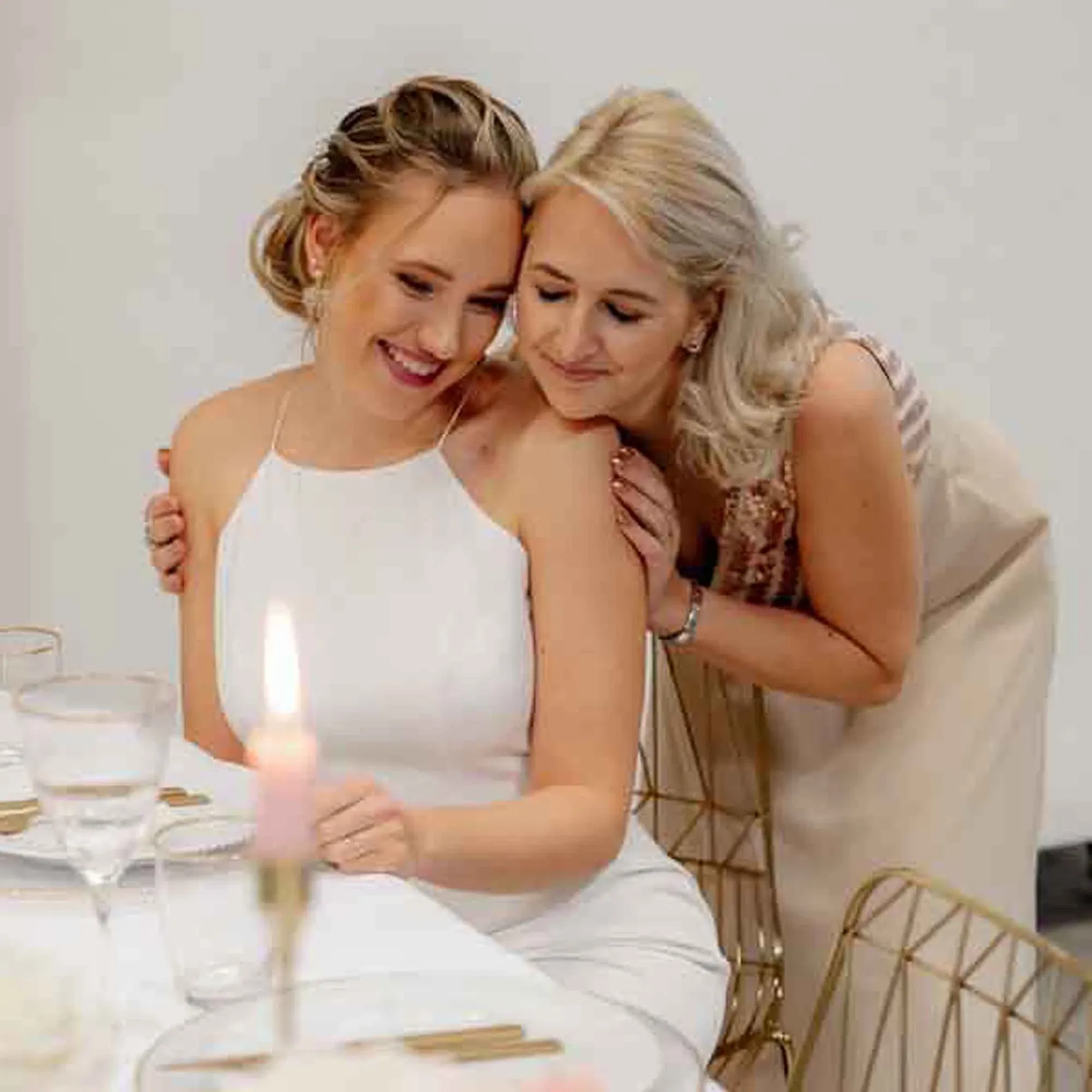 Zwei Frauen in warmem Umarmungs-Moment, Braut am Platz, goldene Stühle und minimalistische Tischdeko im Hintergrund.