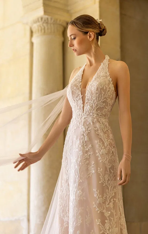 Braut in einem figurbetonten Brautkleid mit tiefem V-Ausschnitt, floraler Spitzenstickerei und fließendem Rock, aufgenommen in einem lichtdurchfluteten, klassischen Säulengang.