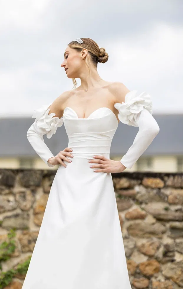 Braut in einem modernen Brautkleid mit herzförmigem Ausschnitt, Off-Shoulder-Ärmeln mit floralen Applikationen und schlichter Linienführung, aufgenommen im Außenbereich vor einer Natursteinmauer.
