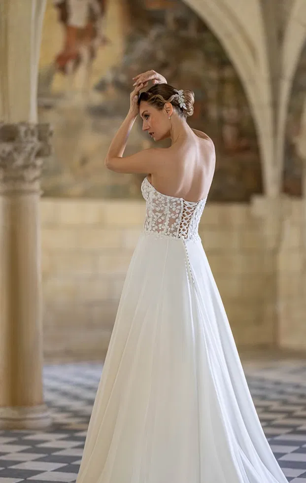 Rückenansicht einer Braut in einem A-Linien-Brautkleid mit transparentem Spitzenoberteil, Schnürung im Rücken und weich fallendem Tüllrock, inszeniert in einem historischen Kreuzgang.