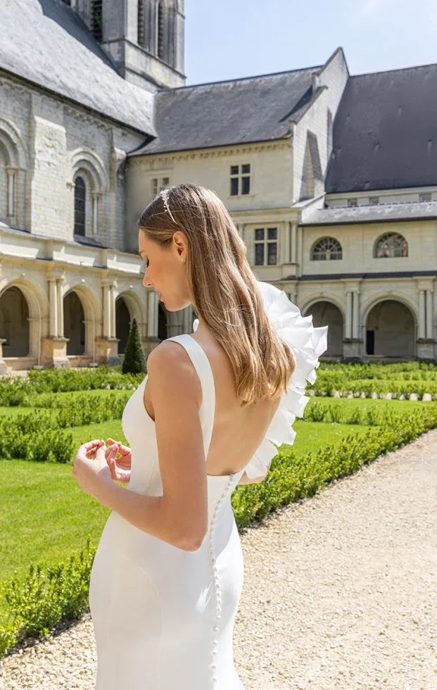 Braut in einem schlichten, modernen Brautkleid mit tiefem Rückenausschnitt und auffälligem, asymmetrischem Volant-Detail an der Schulter, aufgenommen im Garten eines historischen Gebäudes.