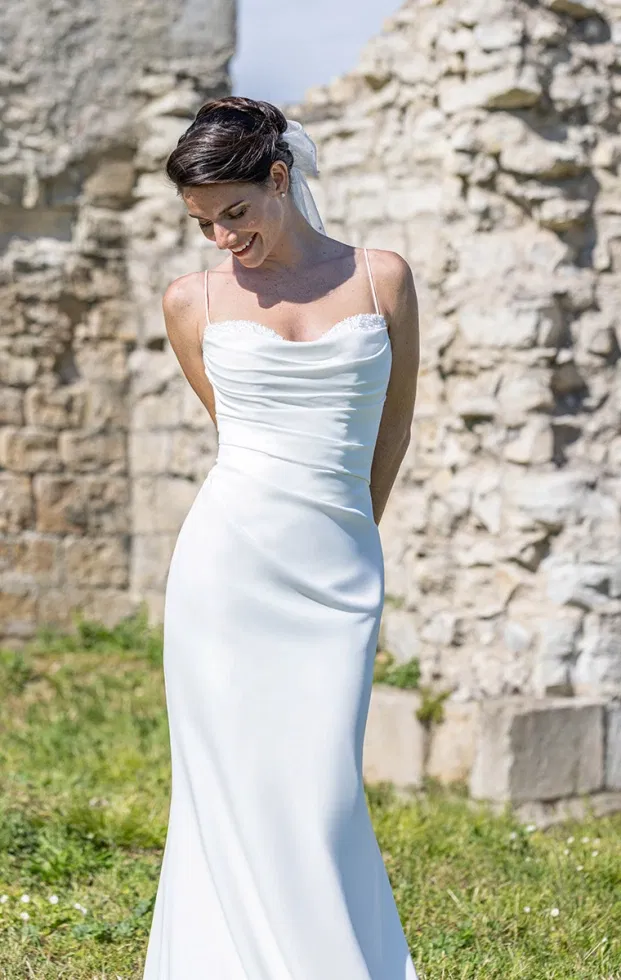 Braut in einem schlichten, figurbetonten Slip-Brautkleid mit zarten Spaghettiträgern und leicht gerafftem Dekolleté, aufgenommen vor einer Natursteinmauer im Freien.
