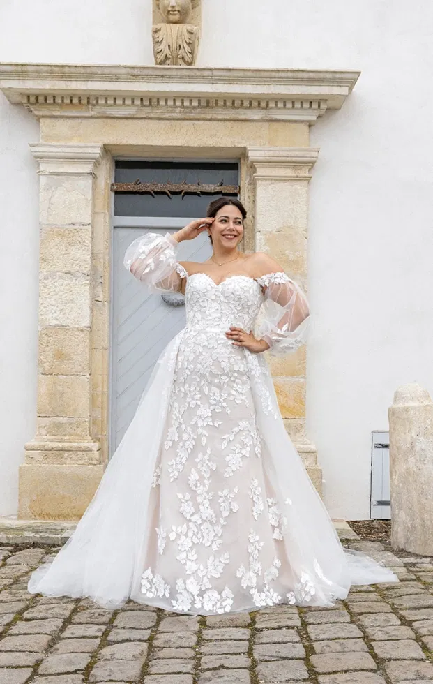 Braut in einem Off-Shoulder-Brautkleid mit transparenten Puffärmeln, floraler 3D-Spitze und weich fallendem Tüllrock, inszeniert vor einem historischen Steineingang.