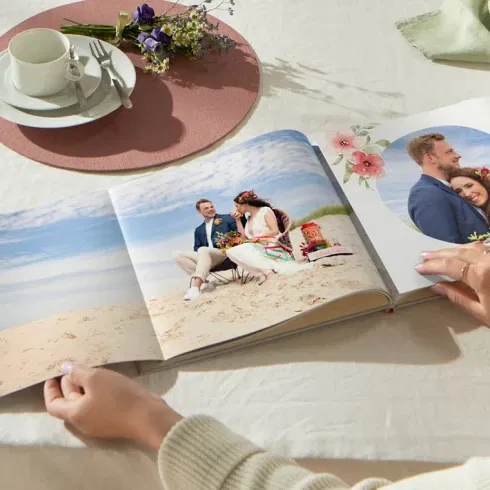 Eine Person blättert in einem Hochzeits-Fotobuch mit Bildern eines Paares am Strand, dekoriert mit floralen Elementen und der Aufschrift „Love“.