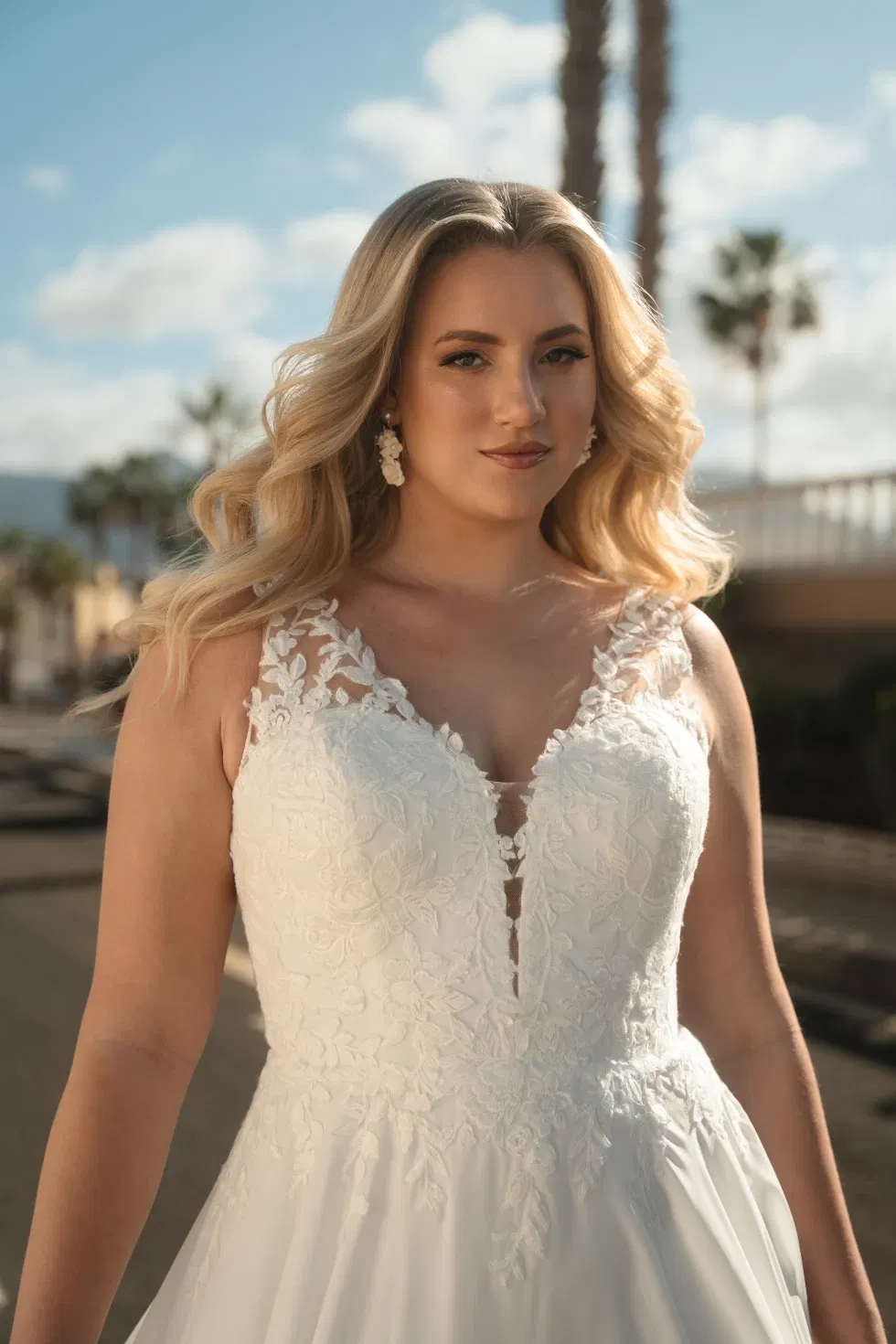 Curvy Braut in einem Spitzen-Brautkleid mit transparentem Dekolleté aus der Madonna-Kollektion von Diane Legrand, aufgenommen im Sonnenlicht mit Palmen im Hintergrund.