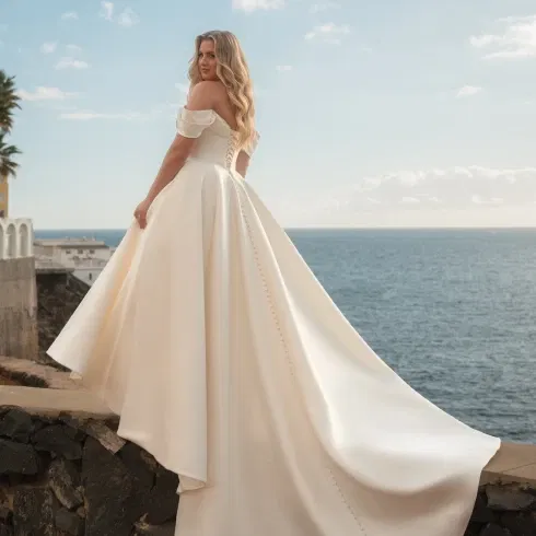 Elegantes Hochzeitskleid im Clean Chic mit abnehmbaren Off-Shoulder-Trägern und langer Schleppe, präsentiert von einer Plus-Size-Braut am Meer.