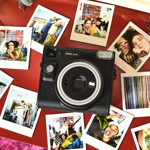 Fujifilm Instax SQ40 Sofortbildkamera liegt auf einem roten Tisch, umgeben von ausgedruckten Sofortbildern, Make-up-Utensilien und Accessoires.