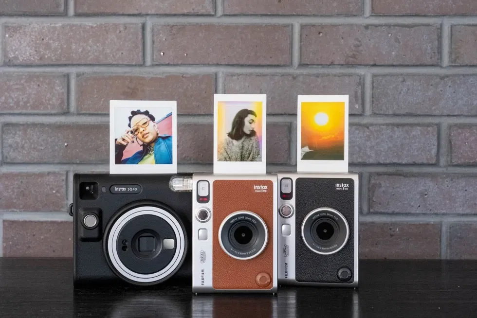 Drei Fujifilm Instax Kameras – die schwarze SQ40 sowie zwei Instax mini Evo Modelle in Braun und Schwarz – stehen nebeneinander vor einer Ziegelwand, jede mit einem Sofortbild im Ausgabeschlitz.