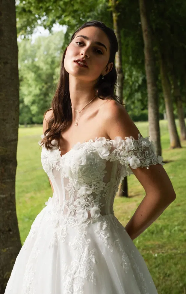 Braut in einem opulenten Anna-Isadora-Brautkleid mit Off-Shoulder-Ärmeln, transparenter Corsage und plastischer Spitzenverzierung, fotografiert im grünen Waldlicht.