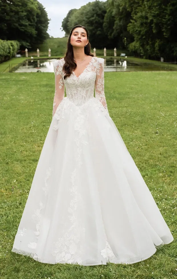 Braut in einem klassischen Anna-Isadora-A-Linien-Brautkleid mit langen Spitzenärmeln, tiefem Ausschnitt und weich fallendem Rock, inszeniert vor einer symmetrischen Gartenkulisse.