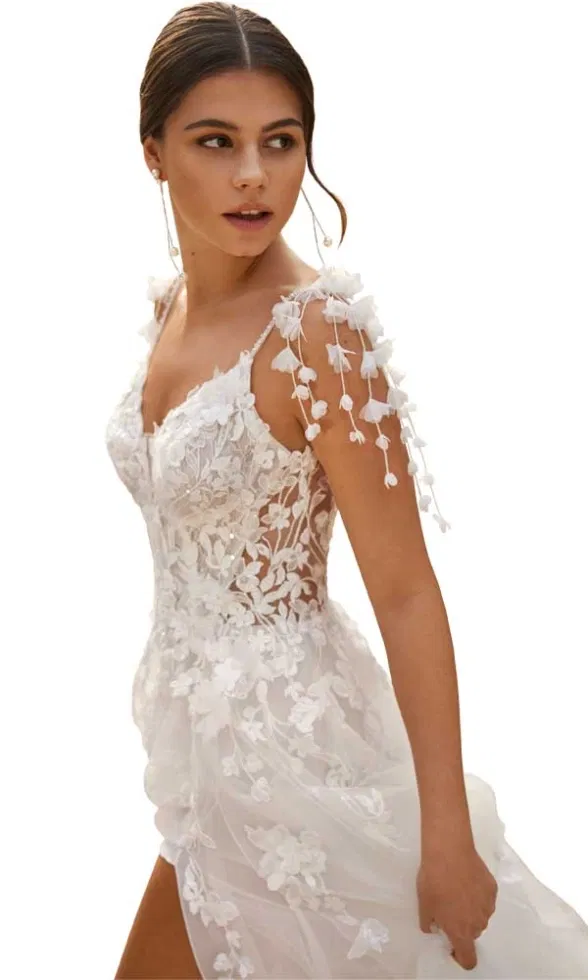 Brautmodell trägt ein figurbetontes Brautkleid mit feiner floraler Spitze, transparenten Trägern und zarten 3D-Blütenapplikationen an der Schulter, elegant inszeniert vor neutralem Hintergrund.