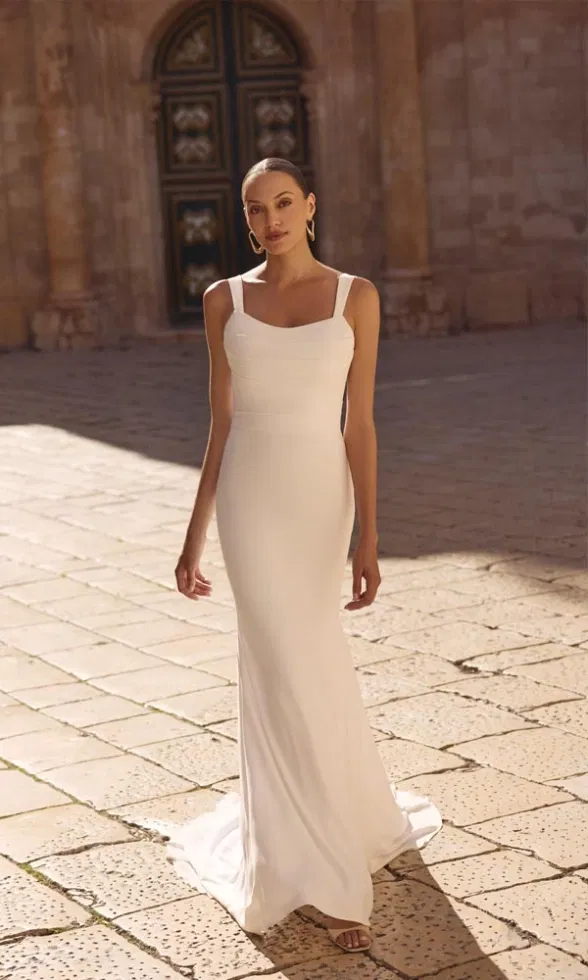 Brautmodell trägt ein puristisches Brautkleid in schmaler Silhouette mit breiten Trägern und geradem Ausschnitt, elegant inszeniert vor historischer Steinarchitektur im warmen Sonnenlicht.