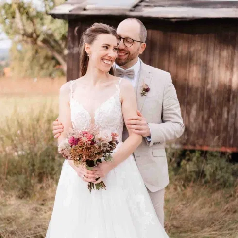 Braut und Bräutigam lächelnd in einer natürlichen Wiesenlandschaft, sie im Spitzenkleid mit Blumenstrauß, er in hellem Anzug.