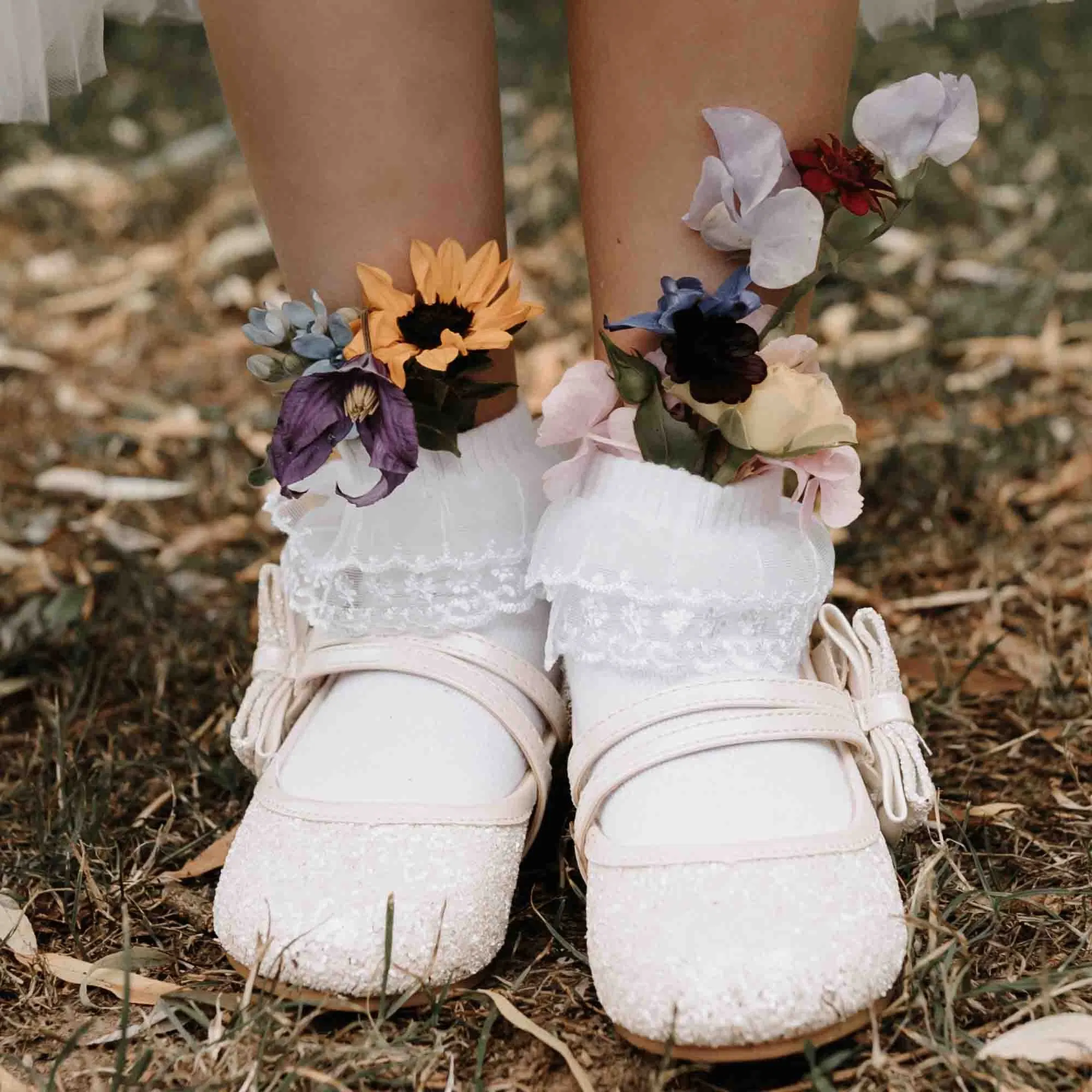 Kinderfüße in weißen Glitzerschuhen mit Rüschen-Söckchen, geschmückt mit bunten Blumen am Knöchel.