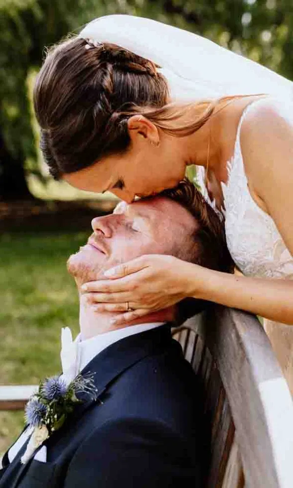 Die Braut küsst sanft die Stirn des Bräutigams, der entspannt auf einer Parkbank sitzt.