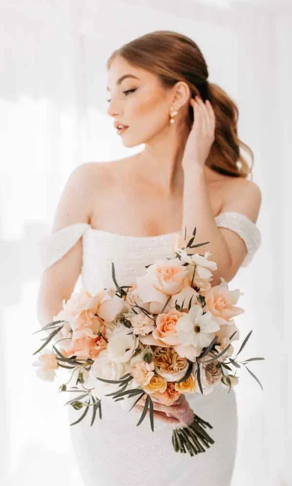 Elegante Braut in einem schulterfreien Kleid hält einen üppigen Brautstrauß in Pfirsich-, Creme- und Rosétönen.