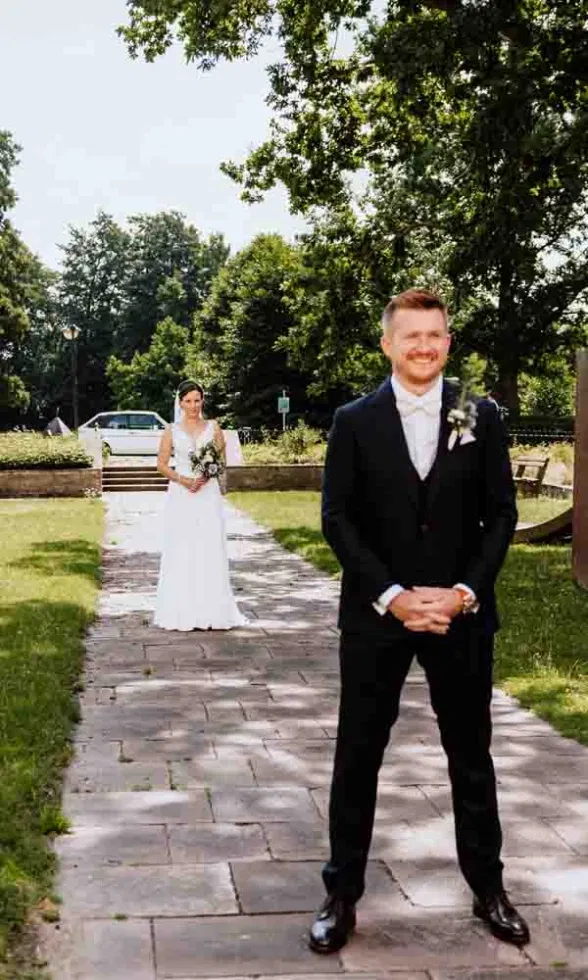 Bräutigam wartet lächelnd, während die Braut für den First Look den Weg entlang auf ihn zugeht.