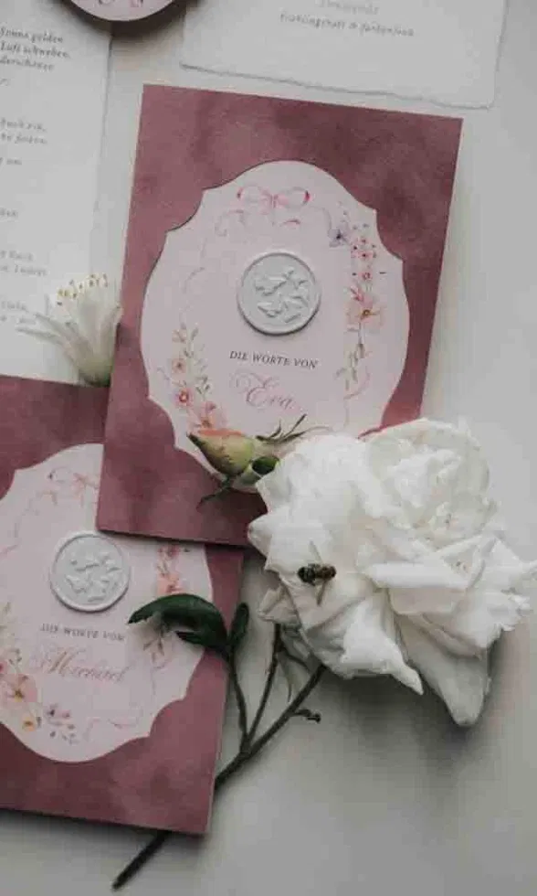 Elegante Hochzeitspapeterie in Rosa mit geprägtem Siegel und floralen Details, dekoriert mit weißen Rosen.