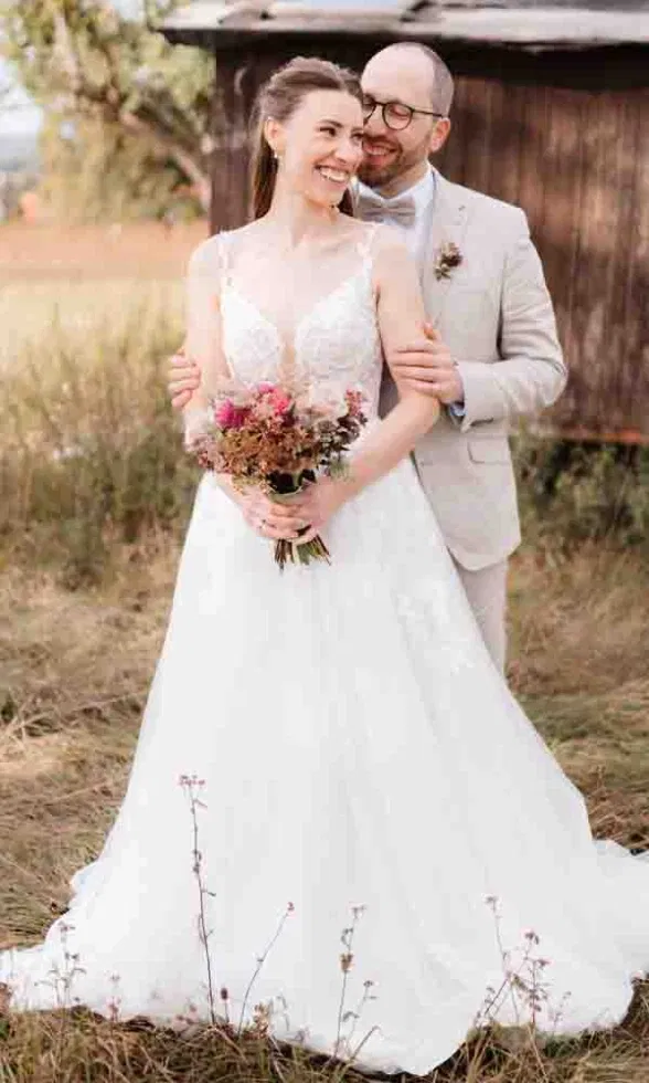 Lächelndes Brautpaar in einer natürlichen Wiesenlandschaft, die Braut im weißen Spitzenkleid mit Blumenstrauß, der Bräutigam umarmt sie von hinten.