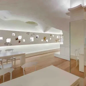 Heller, modern gestalteter Innenraum mit weißen Tischen und Stühlen, hellem Holzfußboden und einer dekorativen Wand mit kleinen beleuchteten Präsentationsfenstern und Vogelmotiven.