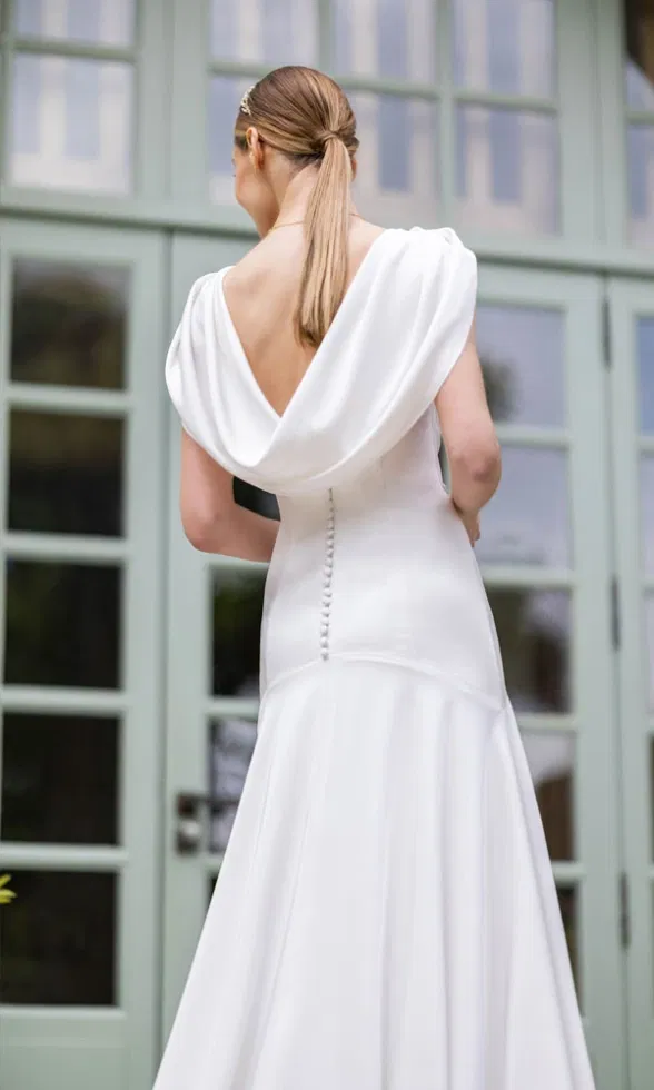 Rückansicht eines Brautkleids mit tiefem Drapé-Rückenausschnitt und Knopfleiste vor großen Glasfenstern.