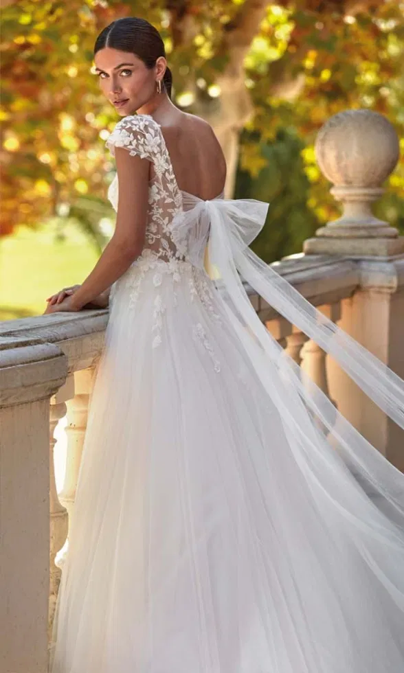 Rückenansicht eines Brautmodells in einem romantischen Brautkleid mit transparentem Spitzenoberteil, floralen Applikationen und großer Tüllschleife am Rücken, inszeniert auf einer Balustrade im warmen Herbstlicht.